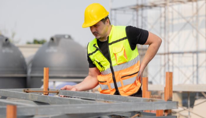 Ein Bauarbeiter mit gelbem Schutzhelm und Warnweste, der sich dem Betrieblichen Gesundheitsmanagement (BGM) verschrieben hat, prüft Metallträger auf einer Baustelle mit großen grauen Tanks und Gerüsten im Hintergrund.