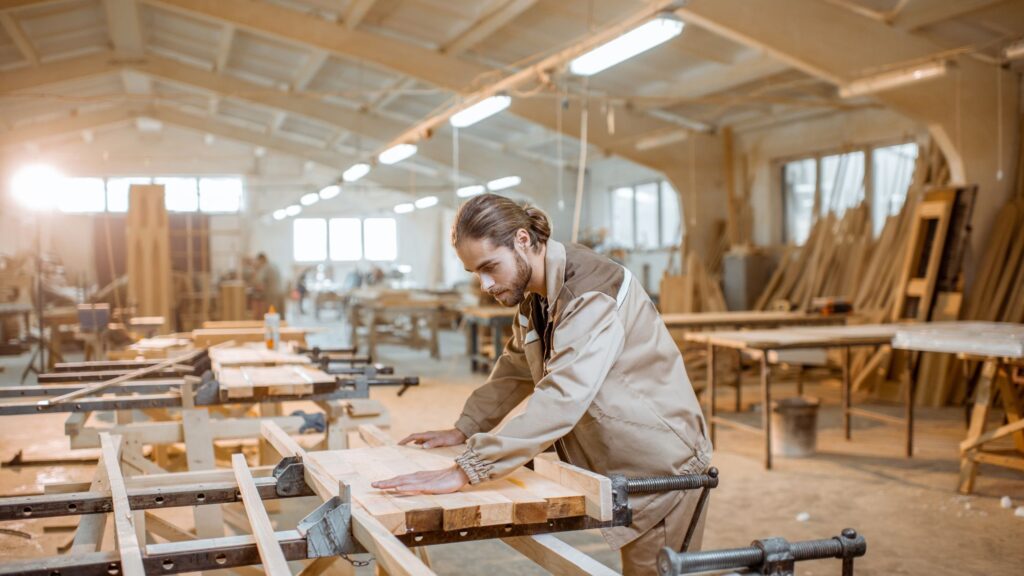 Ein Mann in einem beigen Arbeitsanzug schleift ein Holzbrett in einer geräumigen, gut beleuchteten Werkstatt in Zimmereien, die mit Holzbearbeitungswerkzeugen, Geräten und Holzstapeln gefüllt ist.