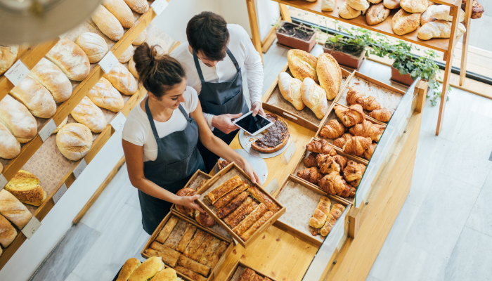 Zwei Personen mit Schürzen stehen in einer Bäckerei, umgeben von einer Auswahl an Brot und Gebäck. Sie scheinen die Artikel in Körben anzuordnen. Im Hintergrund sind Regale voller Brote und weiteres Gebäck zu sehen.