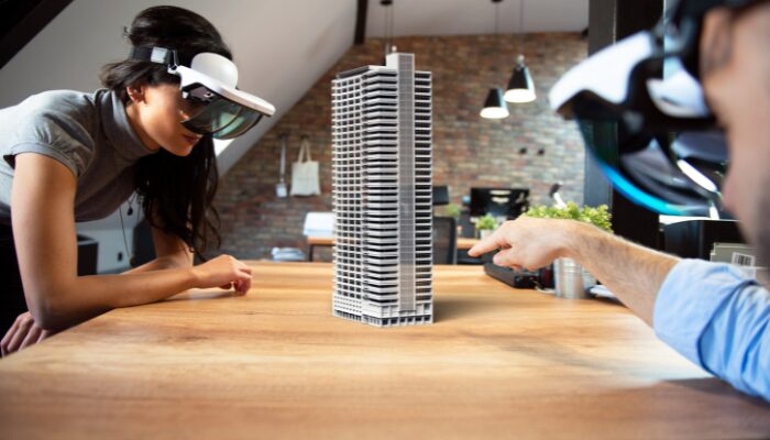 Zwei Personen, die ARvolution-Headsets tragen, interagieren mit einem digitalen 3D-Modell eines modernen Hochhauses, das auf einen Holztisch in einem modernen Büroraum projiziert wird und die Möglichkeiten des Ausbauhandwerks mit erweiterter Realität demonstriert.