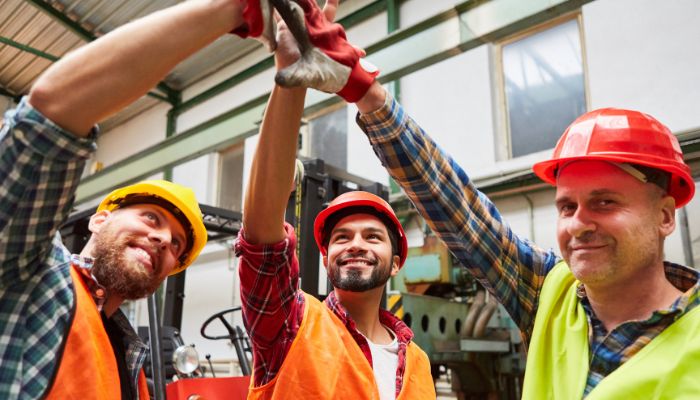 Drei männliche Bauarbeiter in Sicherheitswesten und Helmen, die „Bewährte Methoden im Handwerk“ repräsentieren, lächeln, während sie sich in einem Industriegebäude gegenseitig ein High-Five geben.