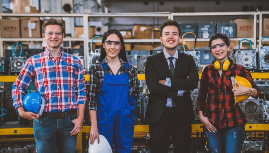Vier junge Erwachsene stehen in einer Fabrik und lächeln in die Kamera. Drei tragen Schutzkleidung und Arbeitskleidung, was modernes Handwerk unterstreicht, während einer im Anzug hohe Arbeitgeberattraktivität widerspiegelt. Dahinter sind Maschinen und Regale mit Kisten zu sehen.