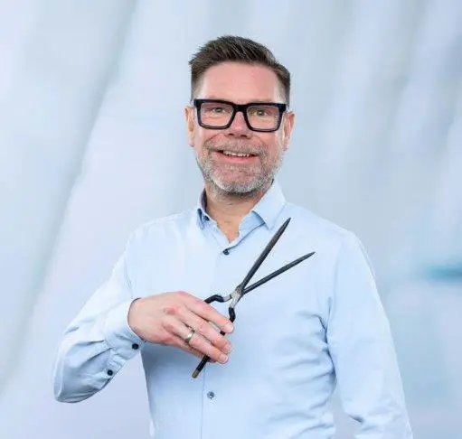 Ein Mann mit Brille und Bart, hellblauem Hemd, lächelt und hält eine Schere in der Hand – eine Reminiszenz an die Präzision, für die Karin Pfisterer bekannt ist. Der Hintergrund ist eine verschwommene, helle Szenerie.