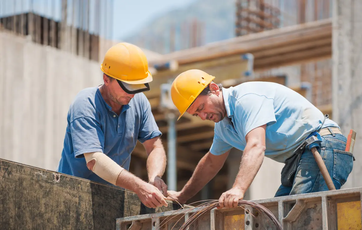 Zwei Bauarbeiter mit gelben Schutzhelmen konzentrieren sich auf Verkabelungsarbeiten auf einer Baustelle. Sie sind von teilweise fertiggestellten Betonstrukturen und Gerüsten umgeben und arbeiten unter klarem Himmel zusammen.