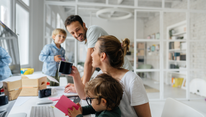 Eine Mutter balanciert Familie und Beruf an ihrem Schreibtisch, lächelnd mit ihrem Kind auf dem Schoß. Ein Mann steht neben einem Jungen im hellen, modernen Home-Office und spiegelt eine echte Work-Life-Balance wider.