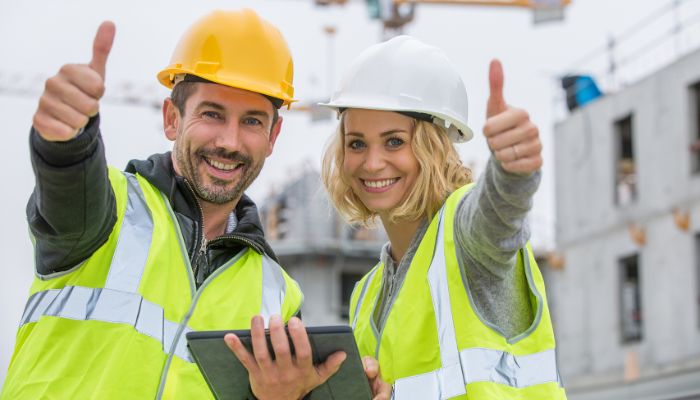 Zwei Bauarbeiter in Sicherheitswesten und Helmen lächeln und zeigen Daumen nach oben, während sie auf einer Baustelle ein Tablet halten, das Digitalisierung und Innovation im modernen Handwerk zeigt, mit Bauaktivitäten im Hintergrund.
