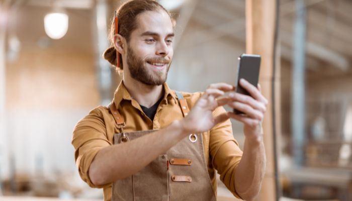 Eine lächelnde Person mit Bart und zurückgebundenen langen Haaren, die eine braune Schürze und ein senffarbenes Hemd trägt, benutzt ein Smartphone in einer hellen, industriellen Werkstatt - ein Beispiel dafür, wie Handwerksbetriebe Social Media nutzen, um sich als attraktive Arbeitgeber zu präsentieren.