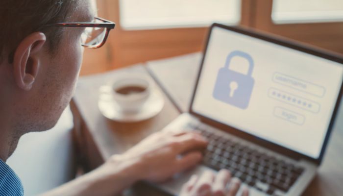 Eine Person benutzt einen Laptop mit einem Anmeldebildschirm, der Felder für Benutzernamen und Kennwort und ein großes Vorhängeschloss-Symbol anzeigt, das auf sichere Daten oder Privatsphäre hinweist. Auf dem Tisch daneben steht eine Tasse Kaffee.
