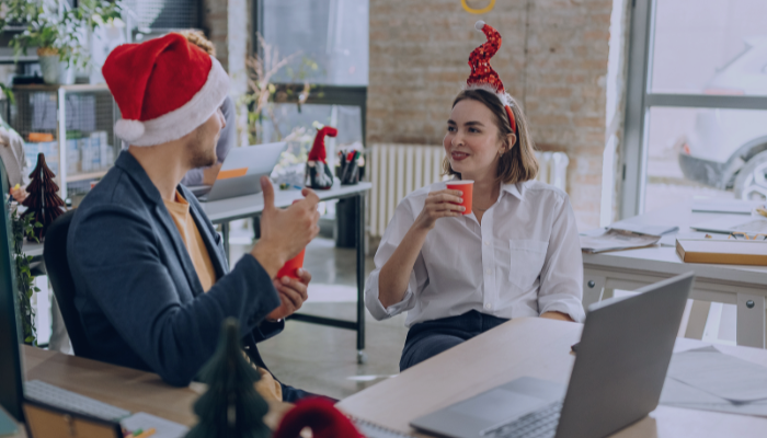 Zwei Kollegen in festlicher Büroatmosphäre, mit Weihnachtsmannmützen und roten Weihnachtsmützen, unterhalten sich bei Getränken gesellig. Laptops und Weihnachtsdekorationen wie Miniatur-Weihnachtsbäume und Lametta schmücken die Schreibtische und unterstreichen ihre zwischenmenschliche Beziehung.