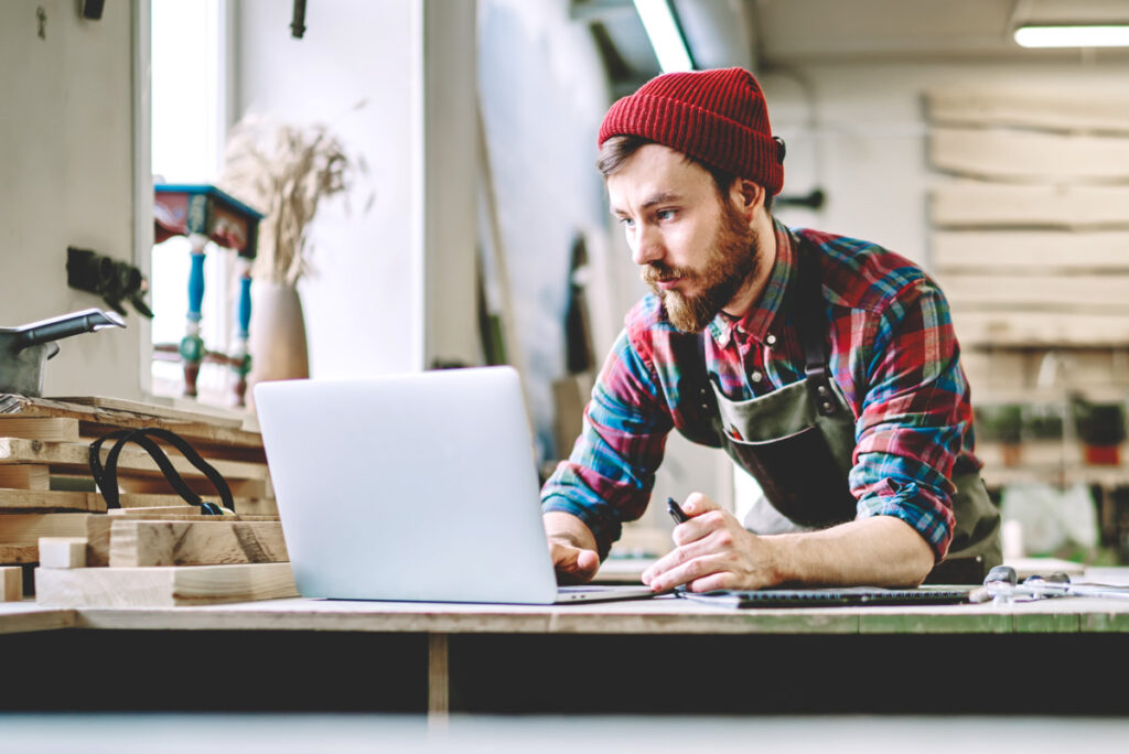 Ein bärtiger Mann mit roter Mütze, kariertem Hemd und Schürze arbeitet in seiner Holzwerkstatt an einem Laptop und verbindet inmitten von Werkzeugen und Holzmaterialien gekonnt traditionelles Handwerk mit Künstlicher Intelligenz.