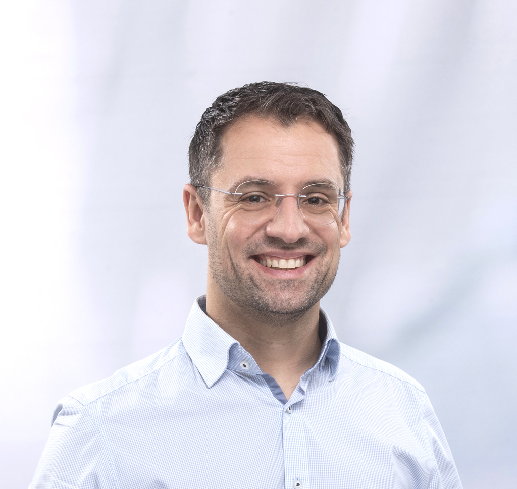 Ein Mann mit kurzen dunklen Haaren und Brille, der ein hellblaues Button-up-Hemd trägt, lächelt in die Kamera vor einem hellen, unscharfen Hintergrund.